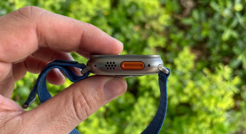 Krupni plan muške ruke koja drži sivi pametni sat Apple Watch Ultra 3 s plavim pletenim remenom i prikazuje narančasti bočni gumb na desnoj strani, s mutnom zelenom vegetacijom u pozadini.