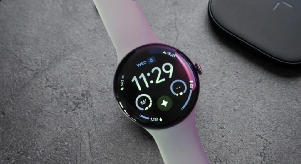 Google Pixel Watch pametni sat u boji Bay s crnim kućištem, na sivoj teksturiranoj površini, s crnim bežičnim punjačem u kutu.